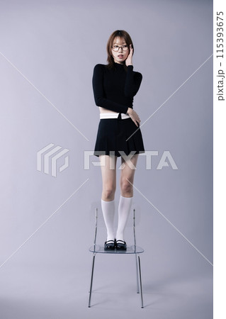 A Korean woman in her 20s posing to touch glasses while standing on a chair wearing a geek look A Korean woman in her 20s posing to touch glasses while standing on a chair wearing a geek look 115393675