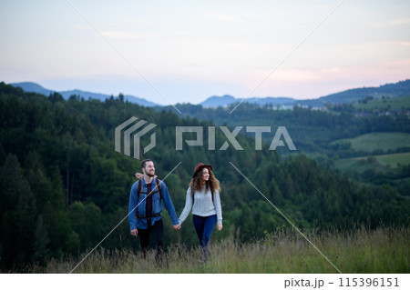 Travellers couple hiking on trail in nature with backpacks, during sunset. Young tourist spending summer vacation oudoors, holding hands. 115396151