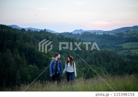 Travellers couple hiking on trail in nature with backpacks, during sunset. Young tourist spending summer vacation oudoors, holding hands. Travellers couple hiking on trail in nature with backpacks, during sunset. Young tourist spending summer vacation oudoors, holding hands. 115396158
