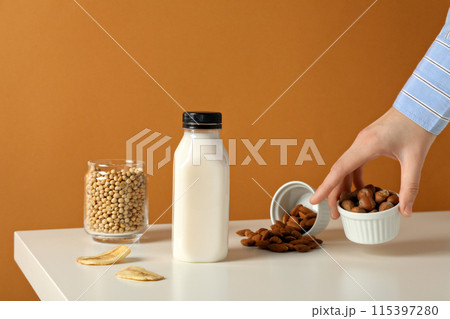 Bottle of milk, hand, nuts in bowls, slices of banana and soy in glass on orange background 115397280