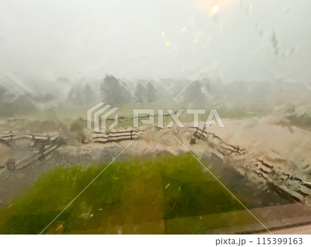 A view through a rain-streaked window capturing a heavy downpour with hail and flash flooding in a suburban backyard. The scene shows blurred fences, trees, and lawn due to the intense rain and hail. 115399163