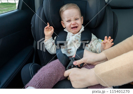 Mother puts her toddler baby into the car seat and fastens seat belt. Baby is sad and crying Mother puts her toddler baby into the car seat and fastens seat belt. Baby is sad and crying 115401270
