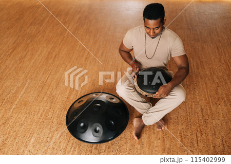 High-angle view of relaxed African man playing traditional ethnic music on glucophone using sticks sitting on floor in lotus pose. Concept of mindfulness, tranquility, harmony, sound therapy. High-angle view of relaxed African man playing traditional ethnic music on glucophone using sticks sitting on floor in lotus pose. Concept of mindfulness, tranquility, harmony, sound therapy. 115402999