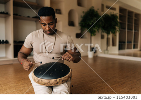 Front view of African man playing ethnic music on tank drum using drumsticks sitting on floor in lotus pose. Serene black male practicing native music drumming instrument playing using sticks. 115403003