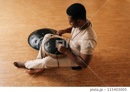 High-angle view of calm African man in trance playing meditative instrument tank drum sitting cross-legged on floor in meditation room. Concept of mindfulness, tranquility, harmony, sound therapy. High-angle view of calm African man in trance playing meditative instrument tank drum sitting cross-legged on floor in meditation room. Concept of mindfulness, tranquility, harmony, sound therapy. 115403005