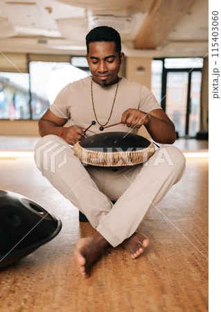 Vertical portrait of calm African man playing traditional ethnic music on glucophone using sticks sitting on floor in lotus pose. Concept of mindfulness, tranquility, harmony, sound therapy. Vertical portrait of calm African man playing traditional ethnic music on glucophone using sticks sitting on floor in lotus pose. Concept of mindfulness, tranquility, harmony, sound therapy. 115403060