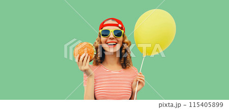 Portrait of cheerful funny young woman with burger fast food and yellow balloon on green background 115405899