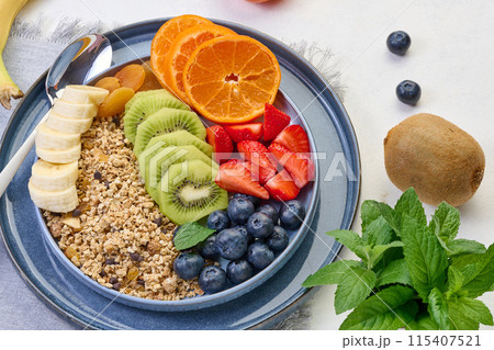 Granola with strawberries, kiwi, banana and blueberries in a round plate on a white table. 115407521