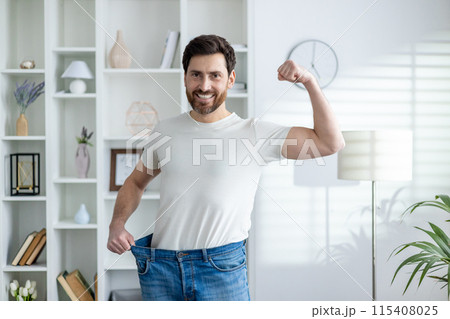 Portrait of a happy and confident young man at home, showing his weight and waist in oversize jeans, showing a sit and victory gesture with his hand. 115408025