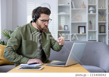 Annoyed young man in headset sitting on sofa at home and talking on video call via laptop, emotionally arguing pointing finger at monitor. 115408042