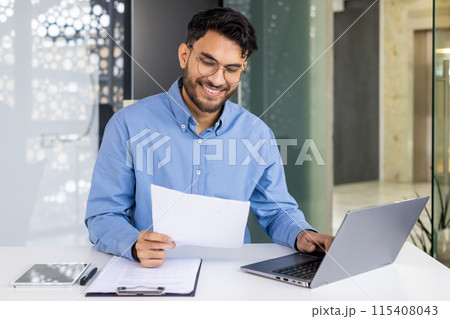 A smiling young Indian man is sitting in the office at a table with a laptop and working with documents, holding papers and typing data on a keyboard. 115408043