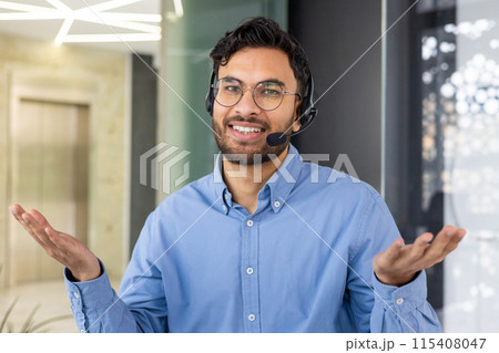 Close-up portrait of a smiling young Indian man in glasses and a blue shirt wearing a headset is in the office, looking at the camera and spreading his hands. Close-up portrait of a smiling young Indian man in glasses and a blue shirt wearing a headset is in the office, looking at the camera and spreading his hands. 115408047