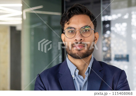 Close-up portrait of a serious young Indian man in a business suit and glasses in the office, looking confidently at the camera. 115408068