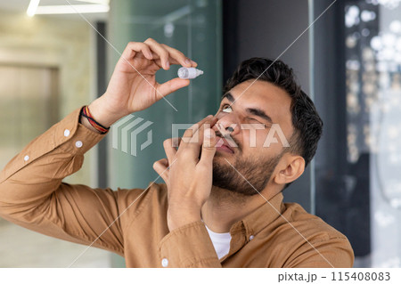 Close-up photo of a young Indian man standing in a modern office and instilling medicine for fatigue and inflammation into his eye. Close-up photo of a young Indian man standing in a modern office and instilling medicine for fatigue and inflammation into his eye. 115408083