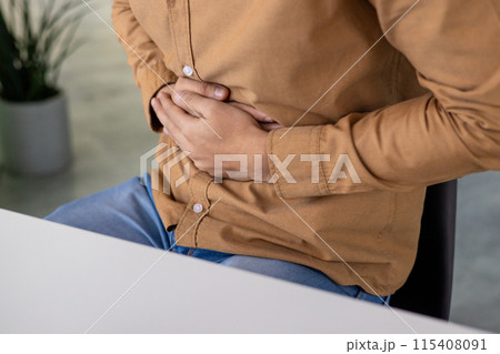 Close-up photo of the hands of a young Hispanic man sitting on a chair at a table and holding his stomach, feeling severe pain and discomfort. 115408091