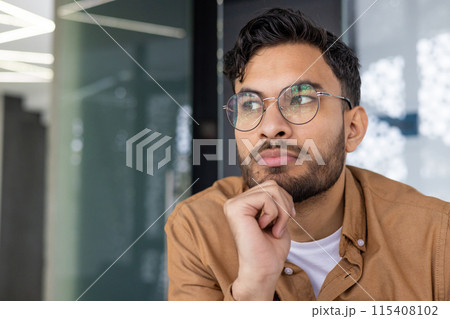 Close-up of a thoughtful young man wearing glasses and looking away, contemplating an idea while sitting indoors. 115408102