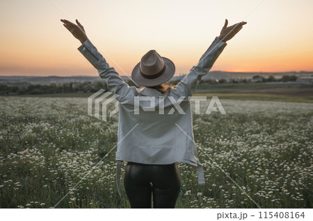A woman wearing a hat stands in a field of flowers, with her arms raised in the air. Concept of freedom and joy, as the woman is celebrating or expressing her happiness. 115408164