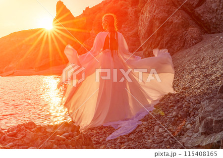 Mysterious woman silhouette long hair walks on the beach ocean water, sea nymph wind listens to the wave. Throws up a long white dress, a divine sunset. Artistic photo from the back without a face 115408165