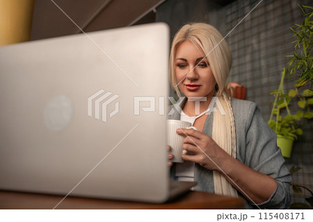 A blonde woman is sitting at a table with a laptop and a white coffee cup. She is wearing a gray jacket and has red lipstick on. The scene suggests a casual and relaxed atmosphere. 115408171