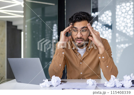 Shocked and upset young Indian man sitting at desk with laptop, crumpled and thrown away paper, tired and frustrated holding his head looking for new ideas. 115408339