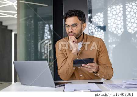 Focused professional working at a modern office desk, analyzing data on a laptop and holding a tablet. Business and technology concepts. 115408361