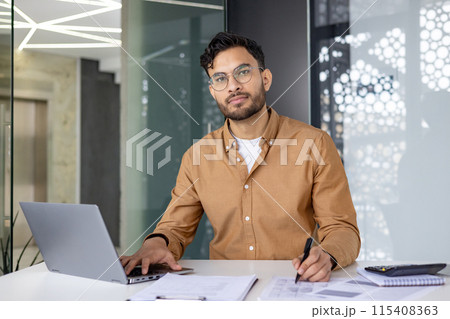Portrait of an Indian young serious man working in the office with a laptop and documents, sitting at a desk, holding a pen and looking at the camera. Portrait of an Indian young serious man working in the office with a laptop and documents, sitting at a desk, holding a pen and looking at the camera. 115408363