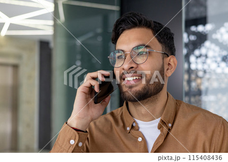 Close-up photo of a young Indian man wearing glasses standing in a modern office center and talking on a mobile phone. Close-up photo of a young Indian man wearing glasses standing in a modern office center and talking on a mobile phone. 115408436