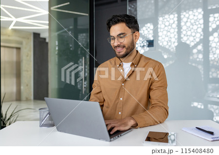 Indian young man studying and working in a modern office, sitting at a desk, smiling and typing on a keyboard with concentration. Indian young man studying and working in a modern office, sitting at a desk, smiling and typing on a keyboard with concentration. 115408480