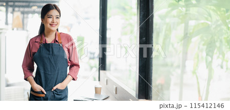 Smiling female barista in apron standing by a window in a modern cafe with a coffee cup and tablet on the counter. 115412146