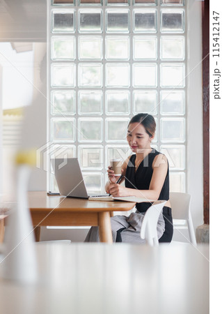 A young professional woman working on her laptop at a modern cafe, holding a coffee cup and smiling, with a glass block wall in the background. 115412147