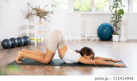 Young woman yoga trainer performing chest-stand pose on black rug 115414948