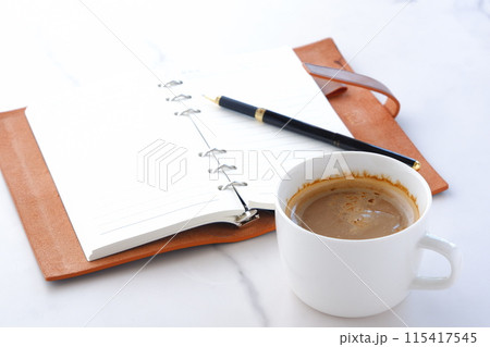 ミルクをたっぷり入れたコーヒーを飲みながら、茶色の革のシステム手帳を開いてデスクワーク中のイメージ ミルクをたっぷり入れたコーヒーを飲みながら、茶色の革のシステム手帳を開いてデスクワーク中のイメージ 115417545