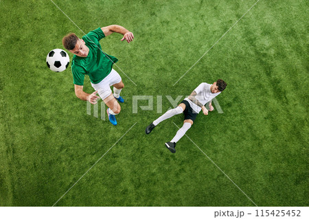 High-angle view. Two soccer players in action on green lush playground. Manin green aims to kick ball, while opponent stays alert and ready to defend. High-angle view. Two soccer players in action on green lush playground. Manin green aims to kick ball, while opponent stays alert and ready to defend. 115425452