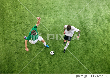 Aerial view. Two players in white and green uniforms battle for the ball on green lush playground. Aerial view. Two players in white and green uniforms battle for the ball on green lush playground. 115425499