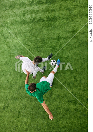 Aerial view. Two young athletic men on field playing football in motion. Soccer players on green lush playground. Aerial view. Two young athletic men on field playing football in motion. Soccer players on green lush playground. 115425540