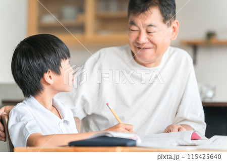 家のダイニングで孫の宿題を教える高齢者男性と小学生の子供 115425640