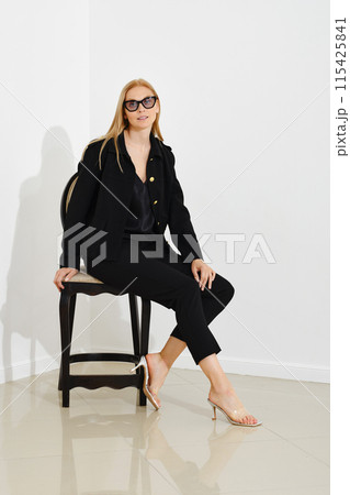 Cute woman in a black jacket and tight trousers sits in profile on a chair against a white wall Cute woman in a black jacket and tight trousers sits in profile on a chair against a white wall 115425841