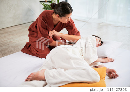Masseuse making thai yoga massage. Woman therapist doing traditional massage treatment for female patient using elbow. Client lying on mat, meditating, enjoying hand massage in calm atmosphere. Masseuse making thai yoga massage. Woman therapist doing traditional massage treatment for female patient using elbow. Client lying on mat, meditating, enjoying hand massage in calm atmosphere. 115427298