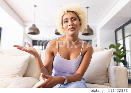 Young biracial woman with curly blonde hair gestures while talking on a video call, seated on a sofa 115432388