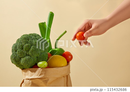 The scene the hand is holding a red cherry tomato in space over yellow background. Beside it is a paper bag, full filled of vegetables contains cauliflower, lemon, leek and so on 115432686