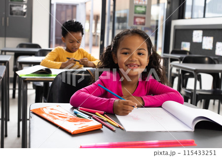 A biracial girl with curly hair smiles while drawing in a school classroom, wearing a pink top 115433251