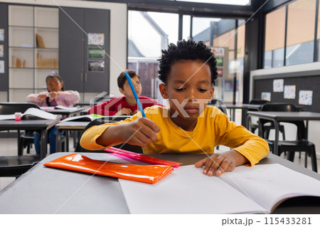 Biracial boy in yellow focuses on writing in a classroom; classmates work in the background Biracial boy in yellow focuses on writing in a classroom; classmates work in the background 115433281