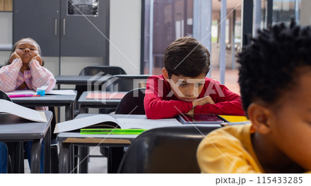 Biracial boy in red looks sad at his desk in a school classroom, with copy space, classmates around 115433285