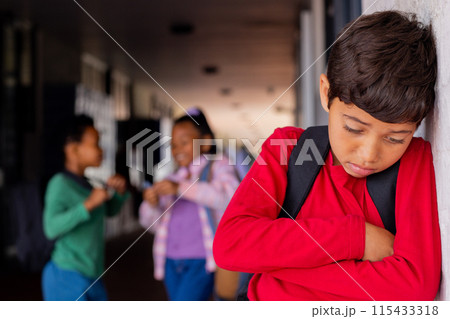 Biracial boy in a red shirt looks upset, leaning against a wall in school, experiencing bullying 115433318