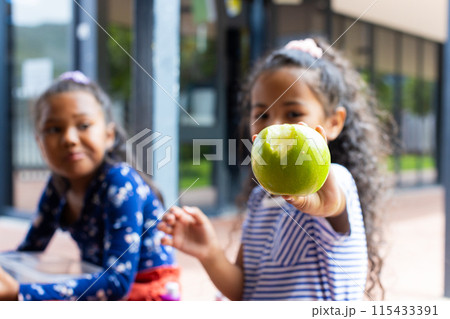 Two biracial girls are sharing a moment in school, one holding a green apple towards the camera Two biracial girls are sharing a moment in school, one holding a green apple towards the camera 115433391