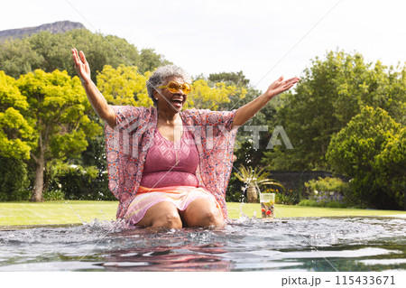 Senior biracial woman enjoys a refreshing moment in a pool, her arms spread wide with joy Senior biracial woman enjoys a refreshing moment in a pool, her arms spread wide with joy 115433671