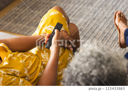 Senior African American woman in a yellow dress holds a phone, seated indoors at home Senior African American woman in a yellow dress holds a phone, seated indoors at home 115433863