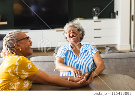 Senior African American woman and senior biracial woman share a laugh together on a couch at home 115433893