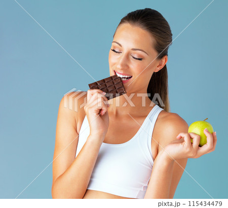 Apple, woman and chocolate choice in studio for health, food and diet plan in isolated blue background. Smile, green fruit and face of young vegetarian person for detox, vegan or decision 115434479