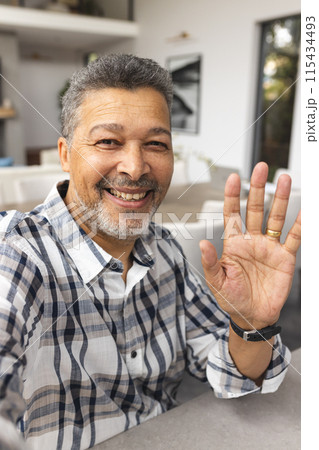 Biracial senior man with gray hair waves at the camera on a video call, sporting a warm smile 115434493